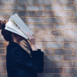 Der Mensch und sein Weib - Eine Frau steht seitlich von einer Backsteinmauer und hat sich mit beiden Händen ein großes Buch auf das Gesicht gelegt.