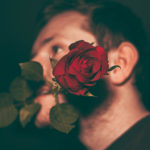 Vereinzelung und die Sehnsucht nach Nähe und freie, sichere Bindungen: Im Hintergrund ist der Kopf eines Mannes von der Seite zu sehen. Quer im Mund hat er den Stiel einer Rose, deren Blüte zentral im Bild im Vordergrund zur Kamera schaut.